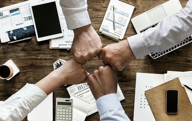 Four hands in 'fist bump' over table with business papers and devices.